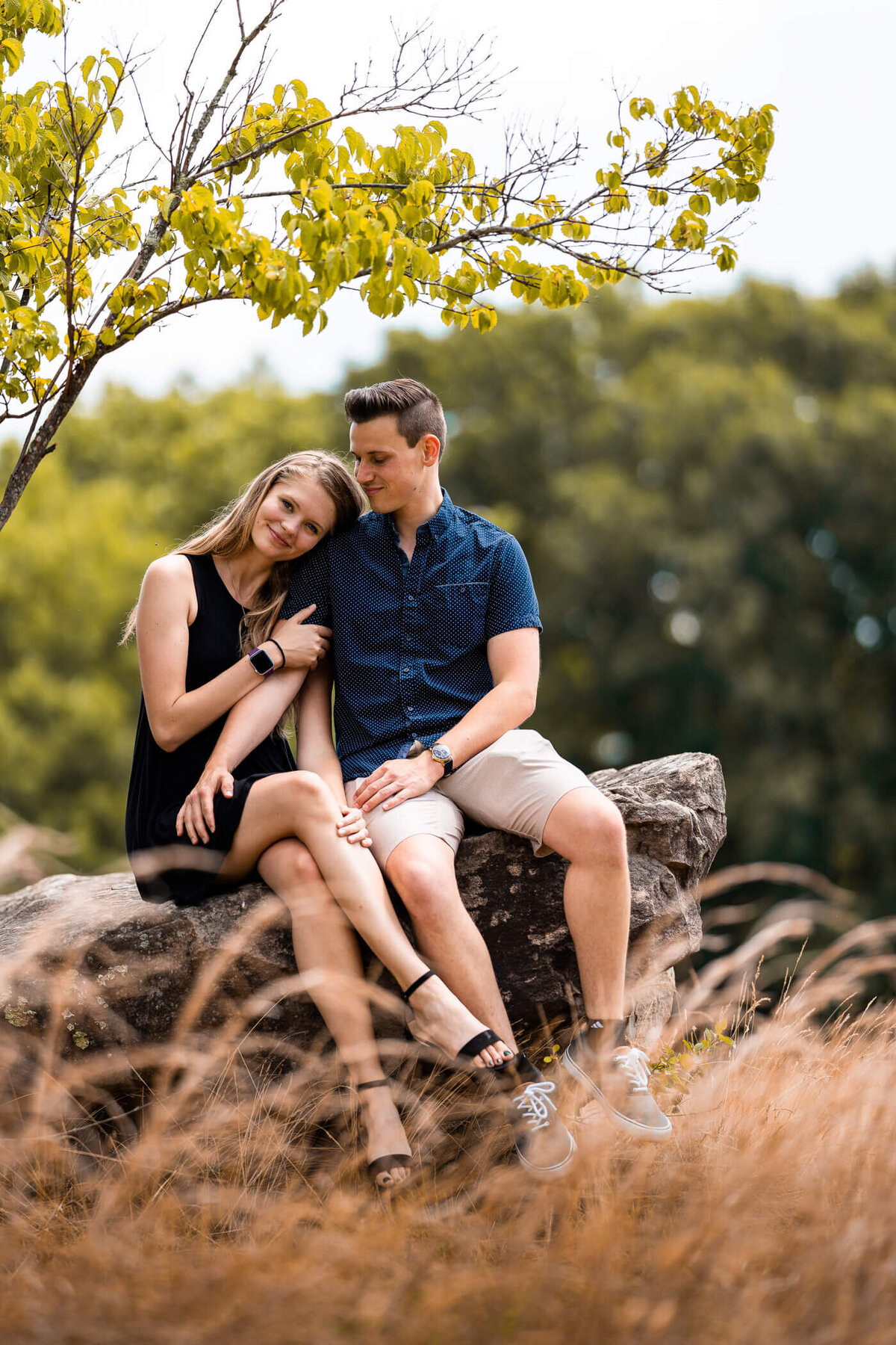 Outdoor portrait of Michael Fricke holding onto his fiancée and smiling while they sit on top of a rock surrounded by a wheat field
