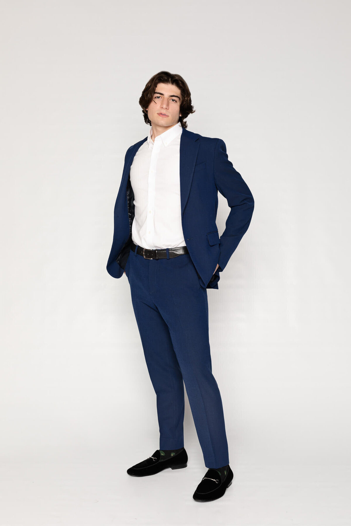A person with medium-length hair is wearing a blue suit, white dress shirt, and black shoes. They are standing confidently against a plain white background.