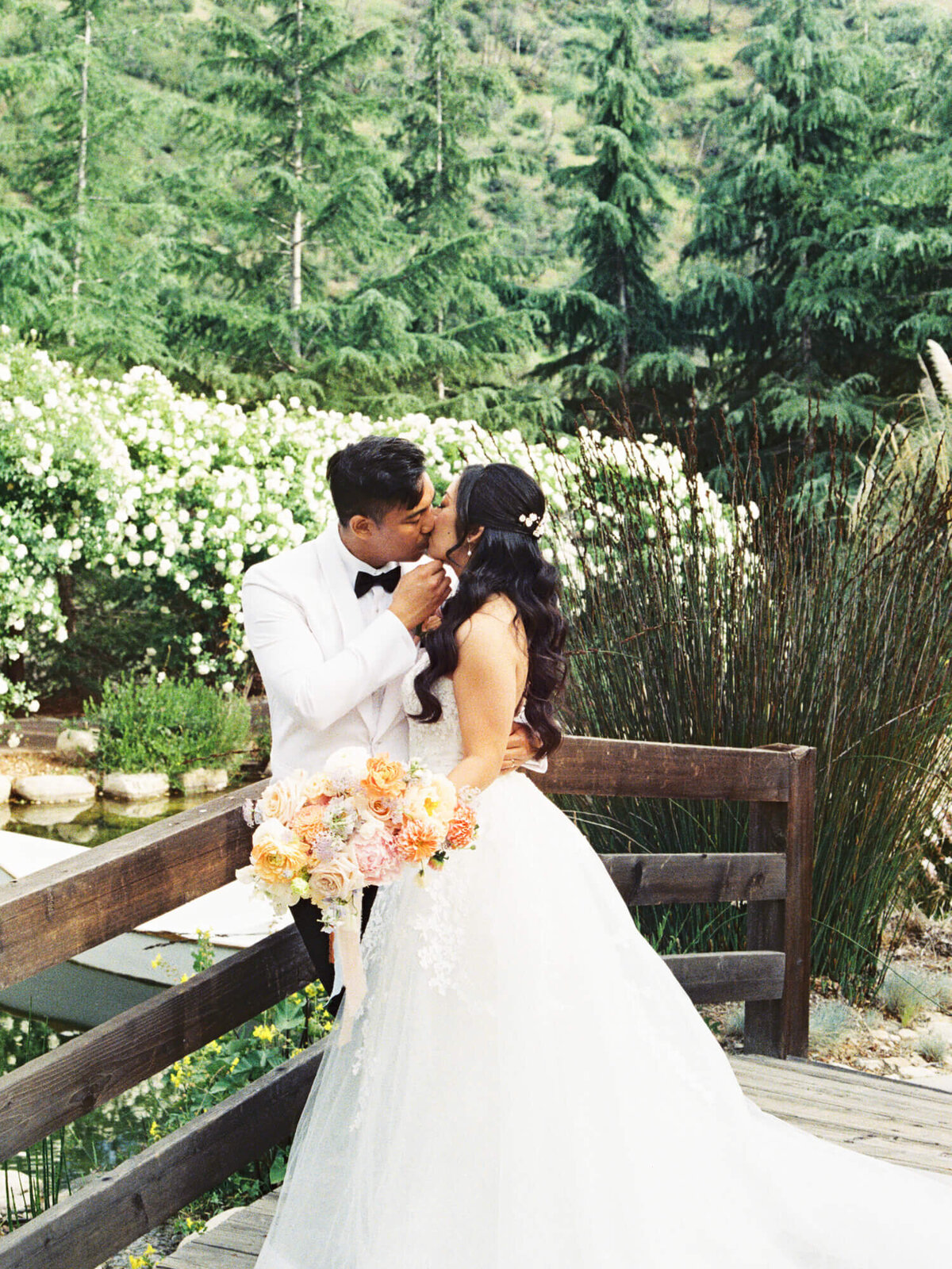 A bride and groom share a kiss on a wooden bridge surrounded by lush greenery and white flowers. The bride holds a bouquet of colorful flowers.