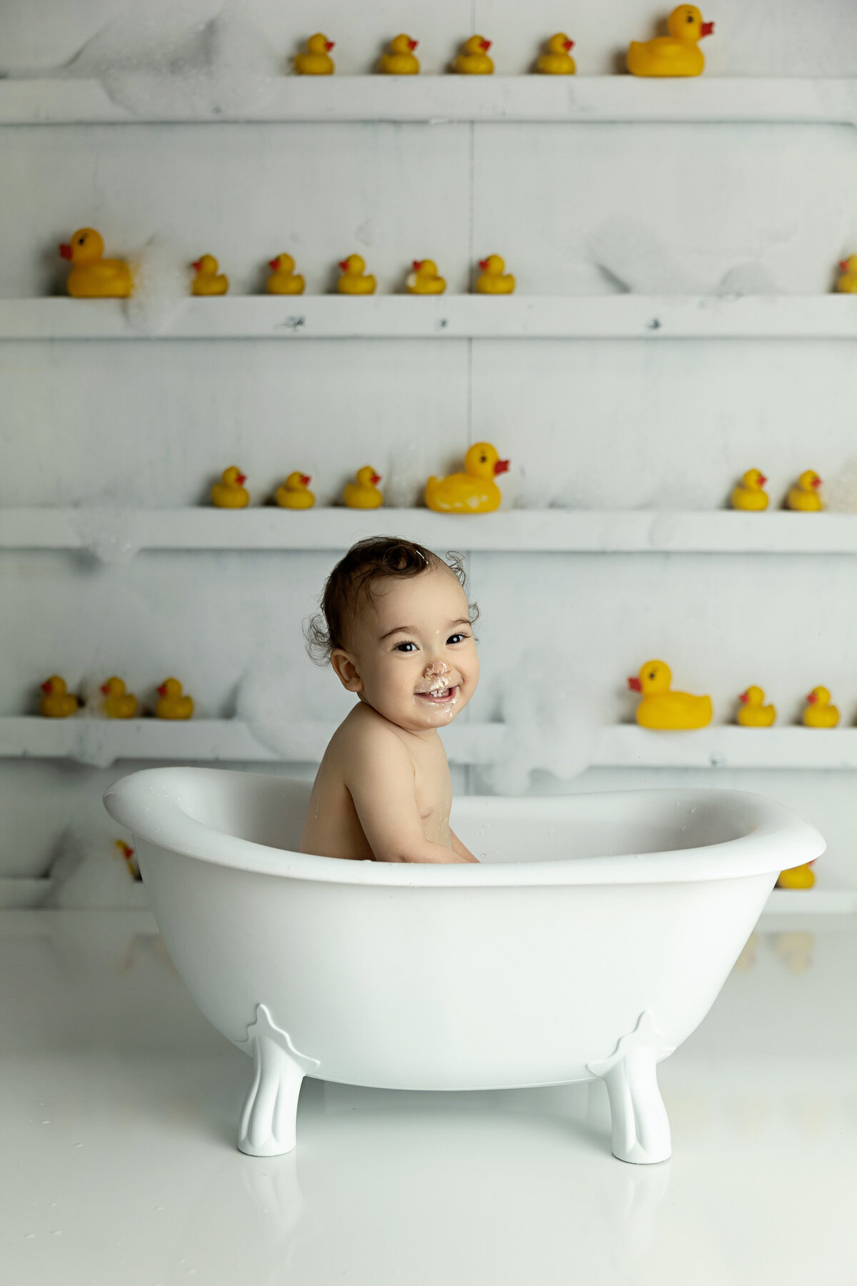 A happy toddler covered in cake sits in a tub with yellow duckies on the wall for a New Jersey Milestone Photographer
