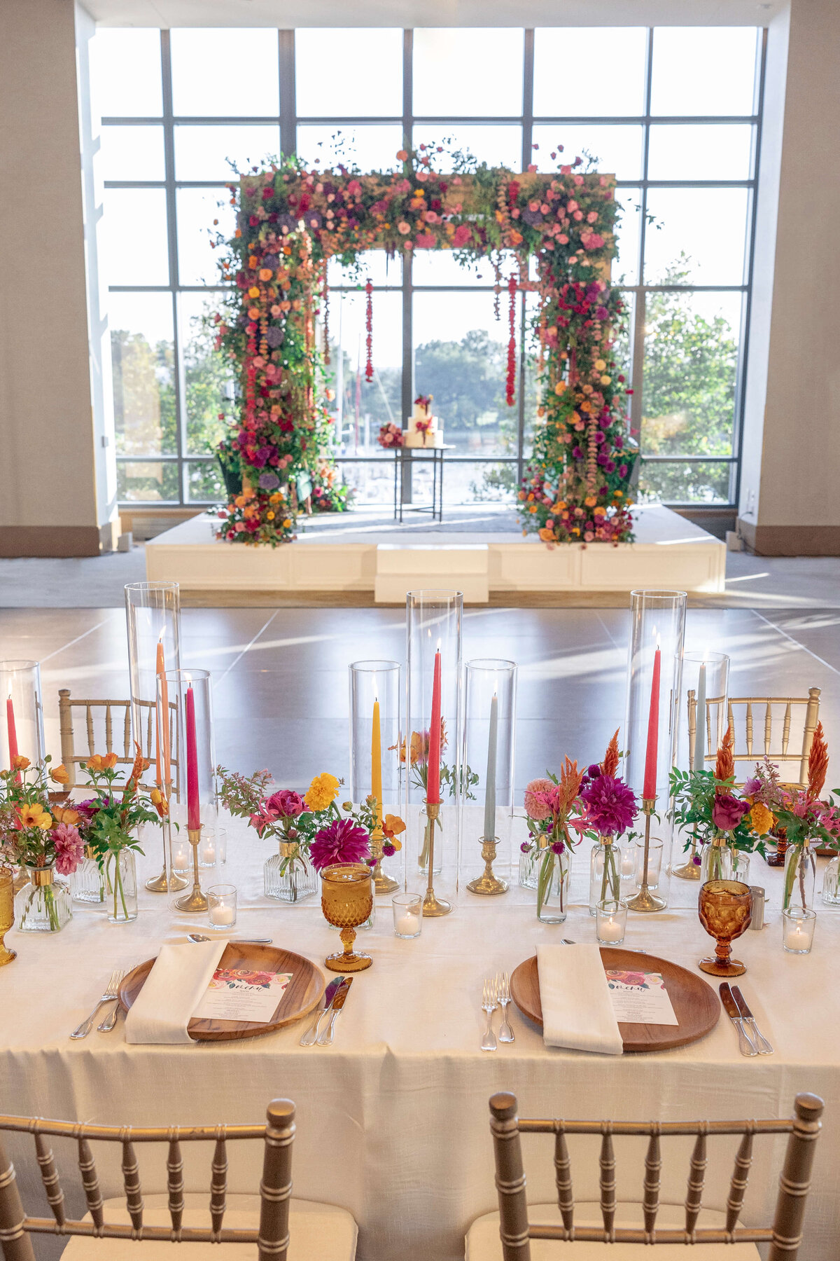 Elegant wedding setup with a floral arch at the altar, featuring colorful flowers. A table in the foreground is adorned with tall candles, floral arrangements, and gold-toned glassware against a bright window backdrop.
