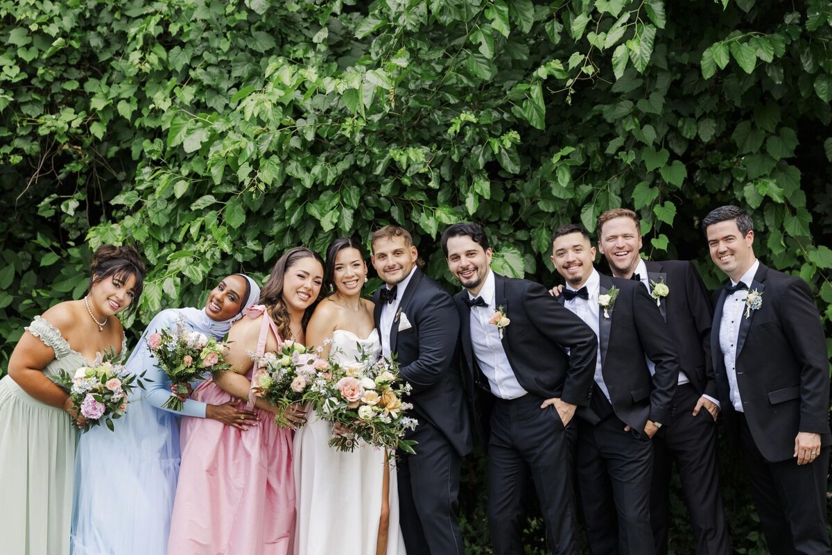 The bride and groom hug the wedding party at The Roundhouse in front of greenery