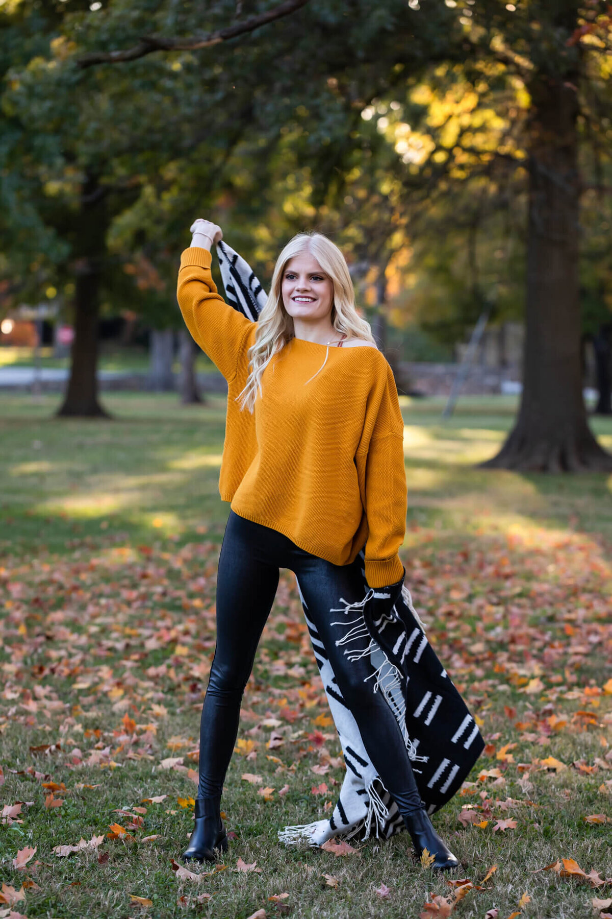 A lovely blonde teen wearing a dark yellow sweater and black leather leggings twirls a black and white blanket behind her in a park covered in Fall leaves. Captured by Springfield, MO senior photographer Dynae Levingston.