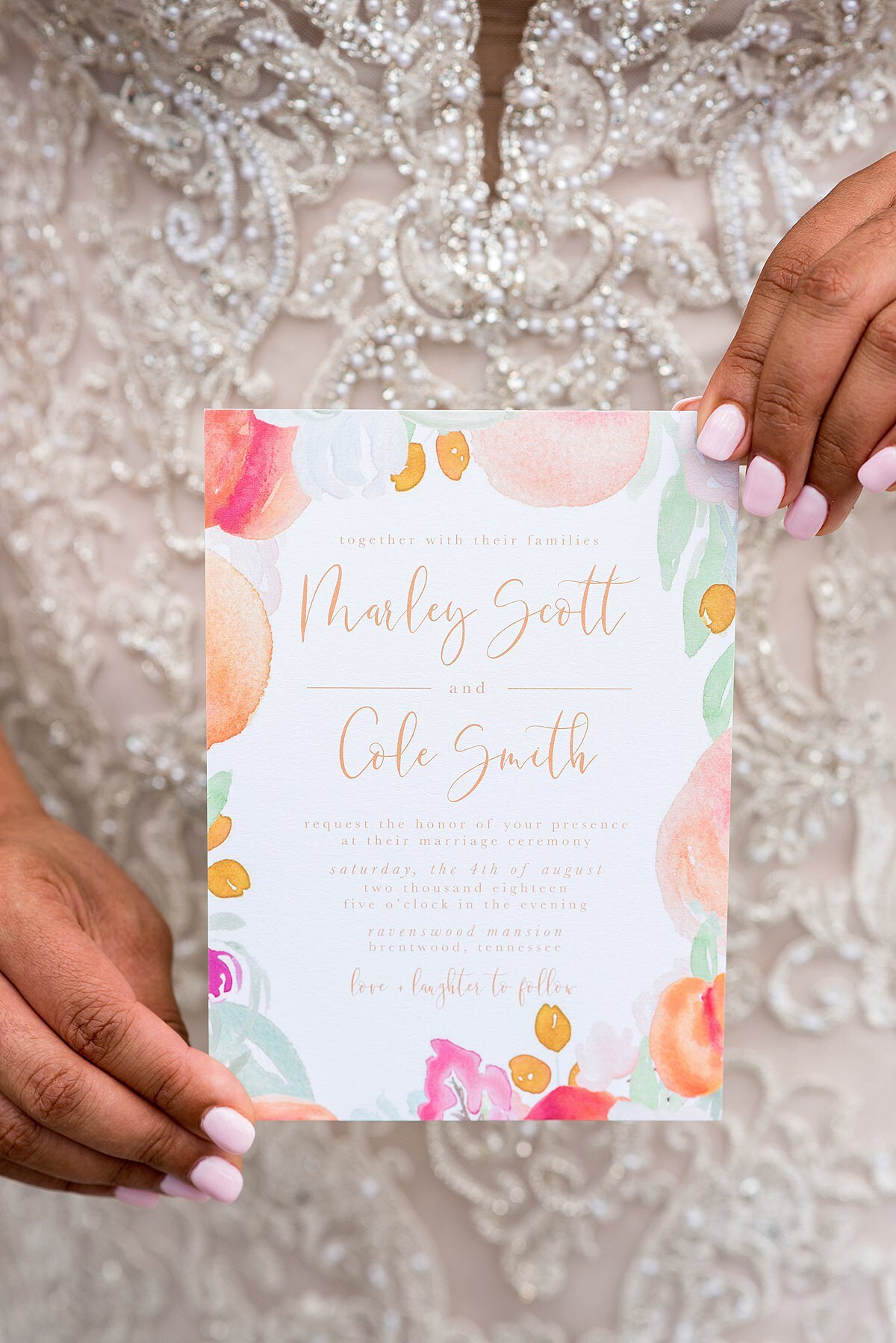 The African American bride, wearing a lace wedding dress, holds a wedding invitation decorated with peaches, pink flowers and green leaves featuring modern calligraphy script at Ravenswood Mansion