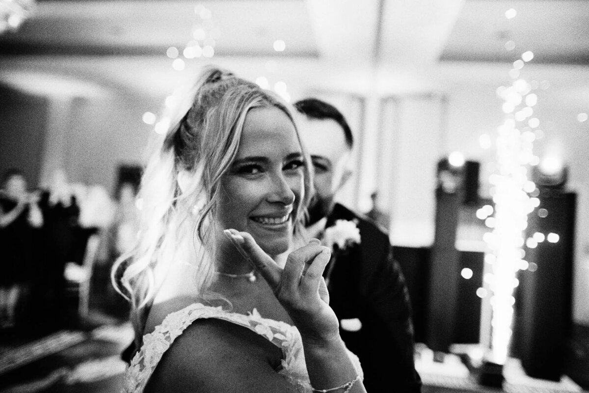 A smiling bride in a wedding dress flashes a peace sign at the camera, with a groom behind her. Captured by a film photographer NJ, the softly lit string lights in the background create a warm, festive atmosphere.