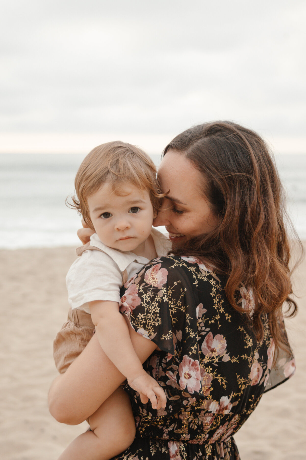 mother snuggles son on beach while son looks at the camera