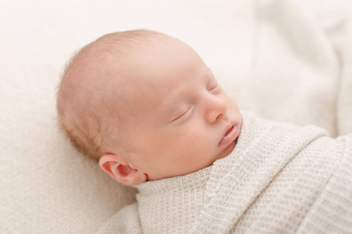 three-quarter profile image of baby's head while baby is sleeping. Baby is swaddled in a beige textured fabric and laying on his back on a beige cozy blanket.