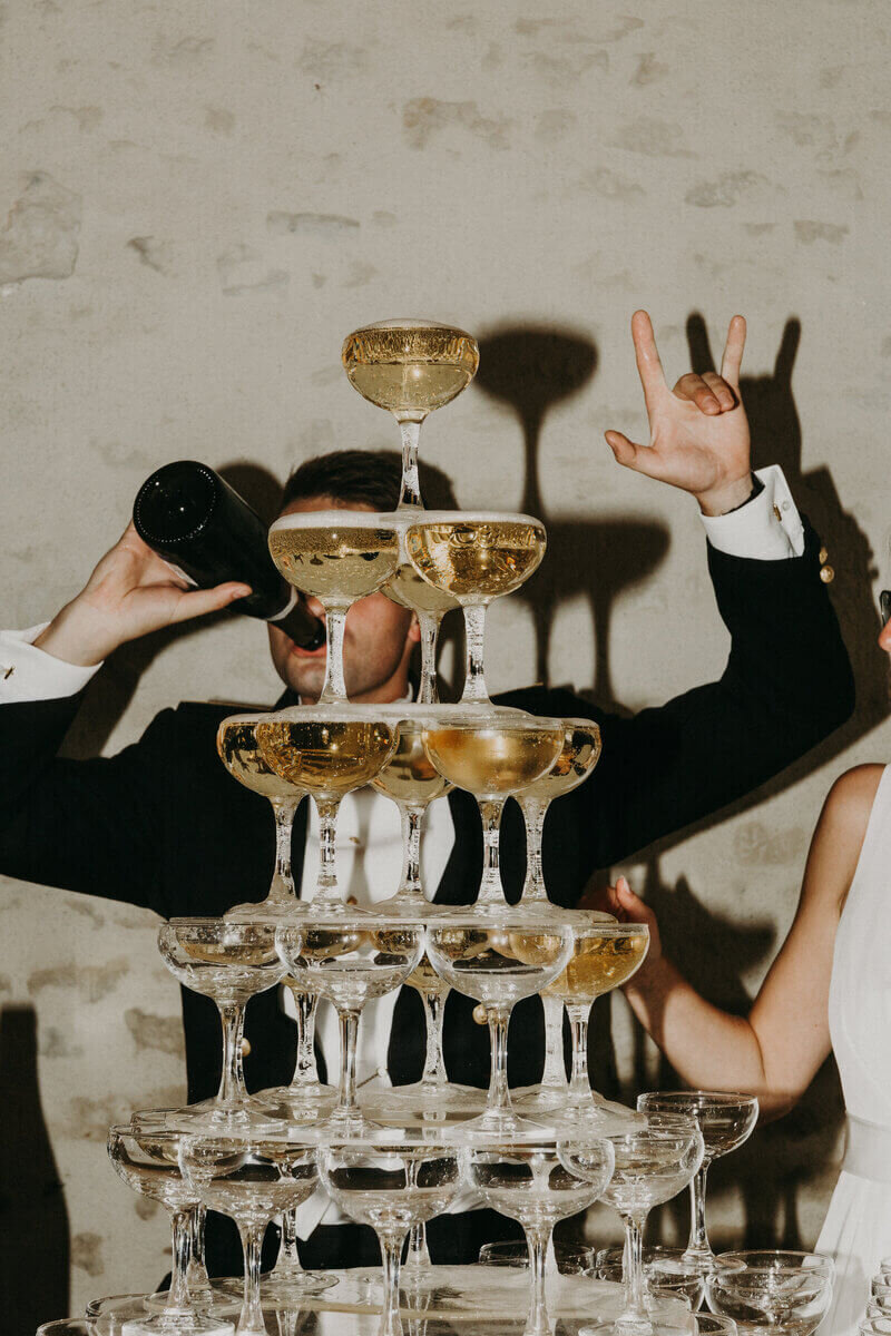 Marié en uniforme buvant le champagne à la bouteille et faisant un signe cool avec son autre main, caché derrière une structure de coupes de champagne. Photo capturée par Laura Termeau photographie.