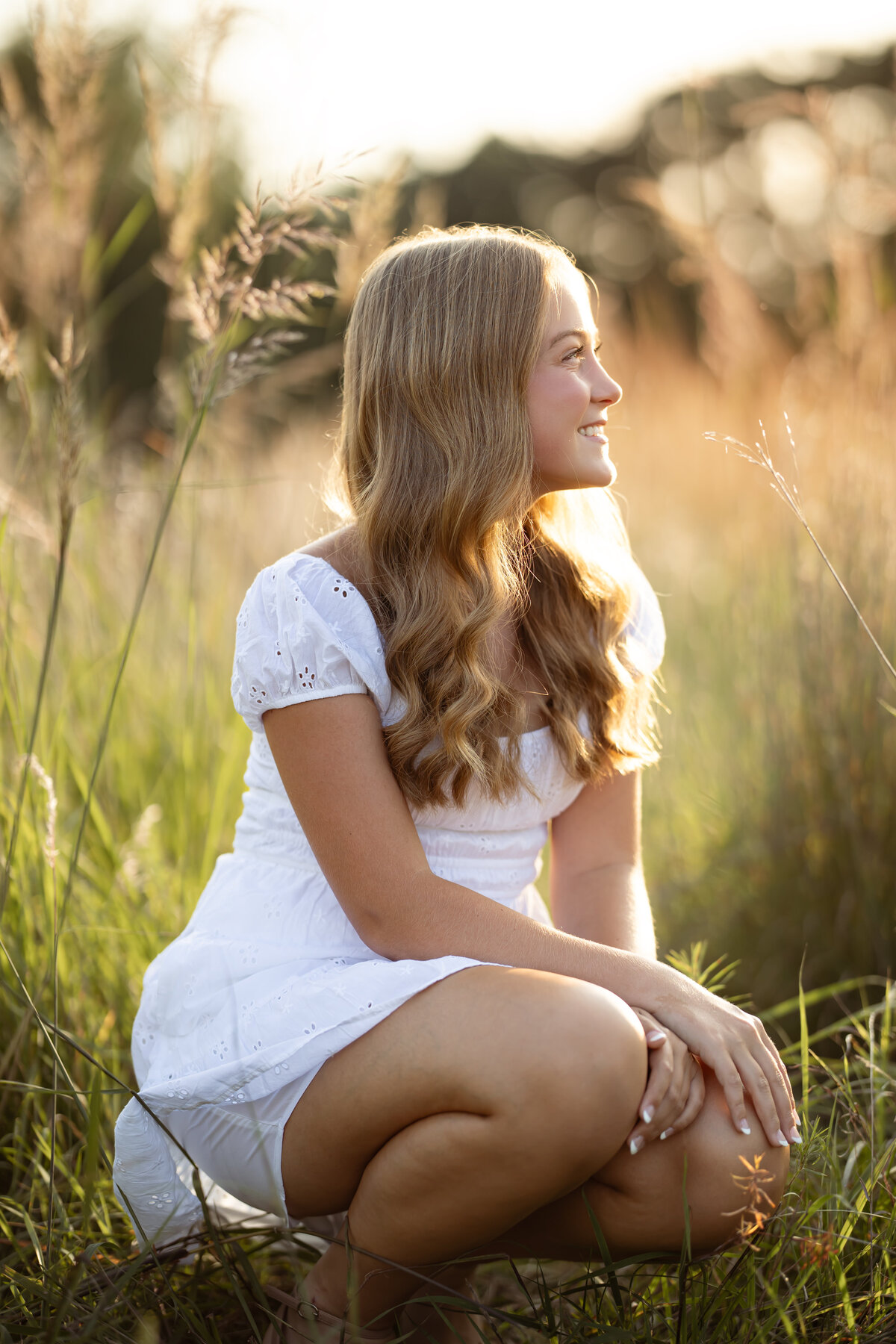 a young woman poses for senior portraits in a field in Eden Prairie, MN. she wears a white dress and kneels, looking away from the camera