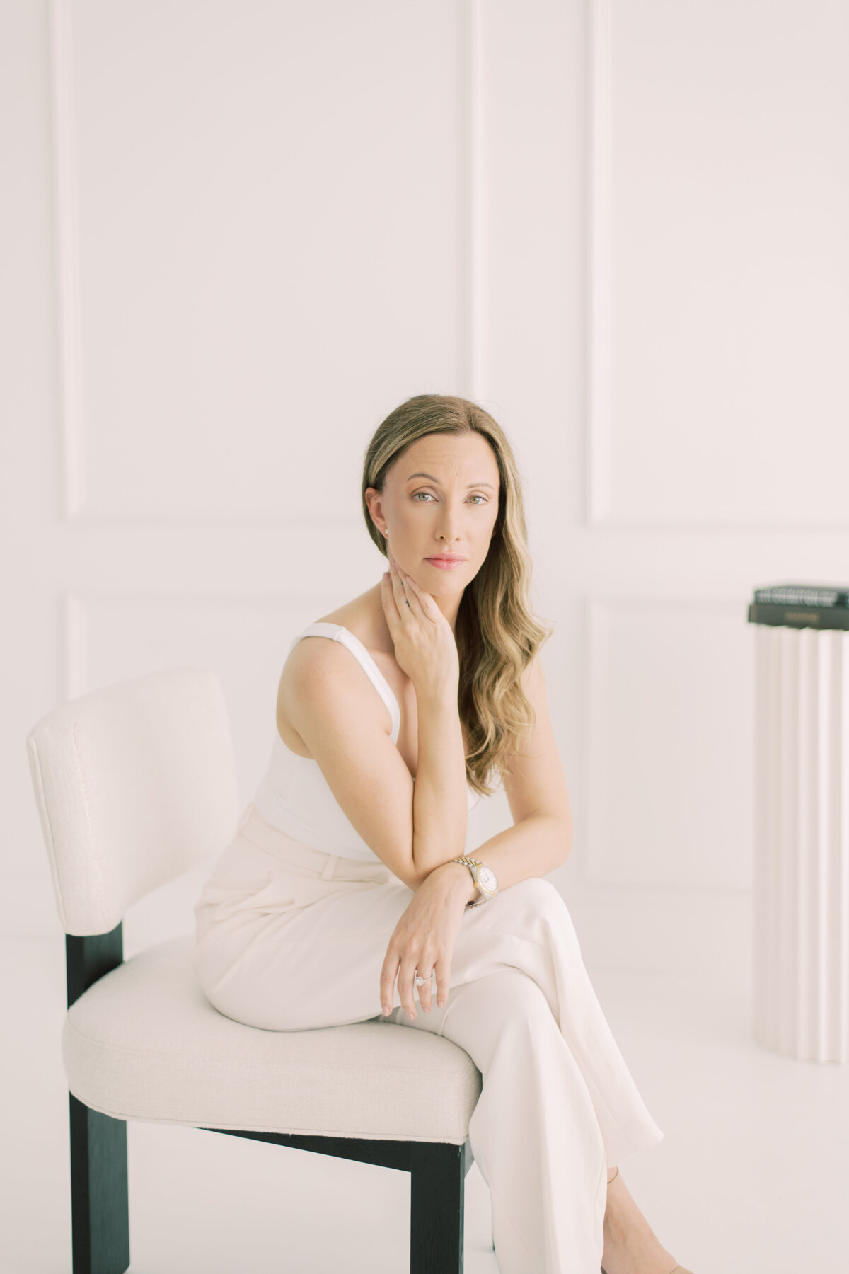 Seated woman in white, softly posed with hand under chin, minimalist studio backdrop
