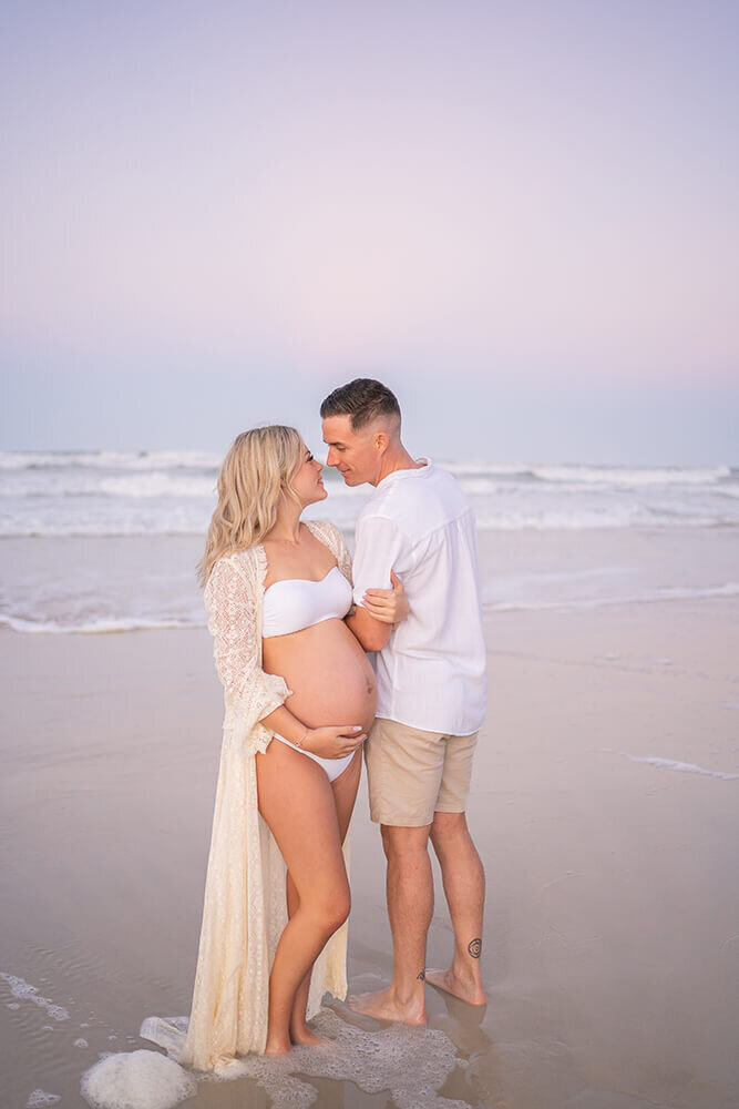 couple posing for pregnancy shoot on the beach pink sunset in the background