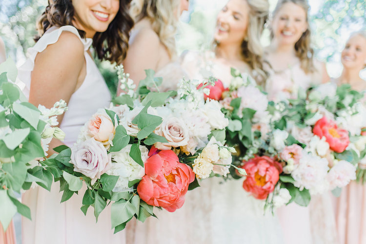 A bride and four bridesmaids in taupe dresses hold their vibrant coral and white bouquets together at a wedding at Chateau Montebello in Quebec