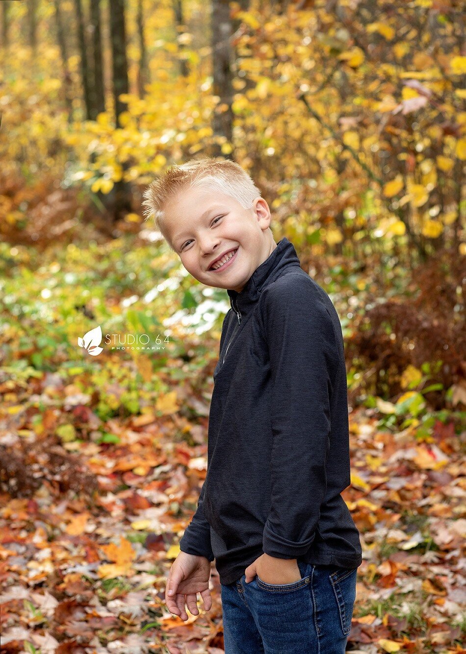 A young boy smiles candidly outdoors with the fall colors surrounding him.