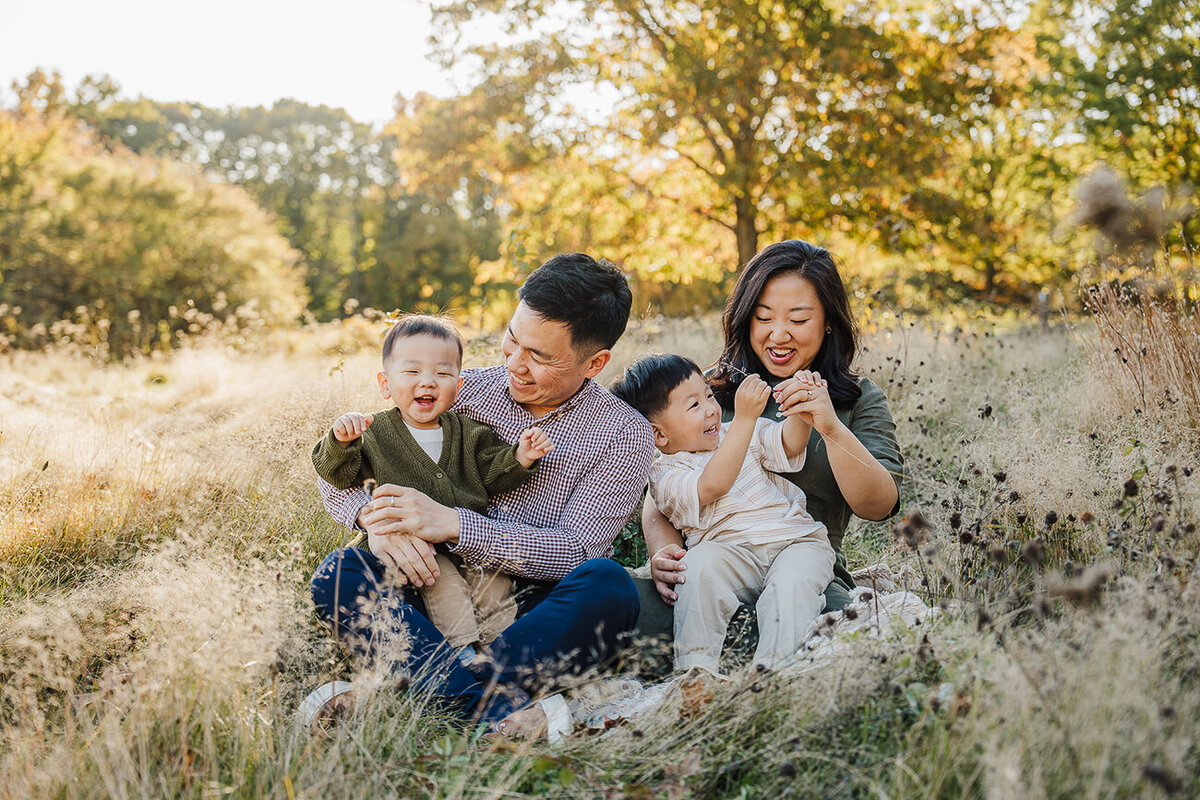 family sits in golden grass and laughs with two toddler sons during photoshoot in boston