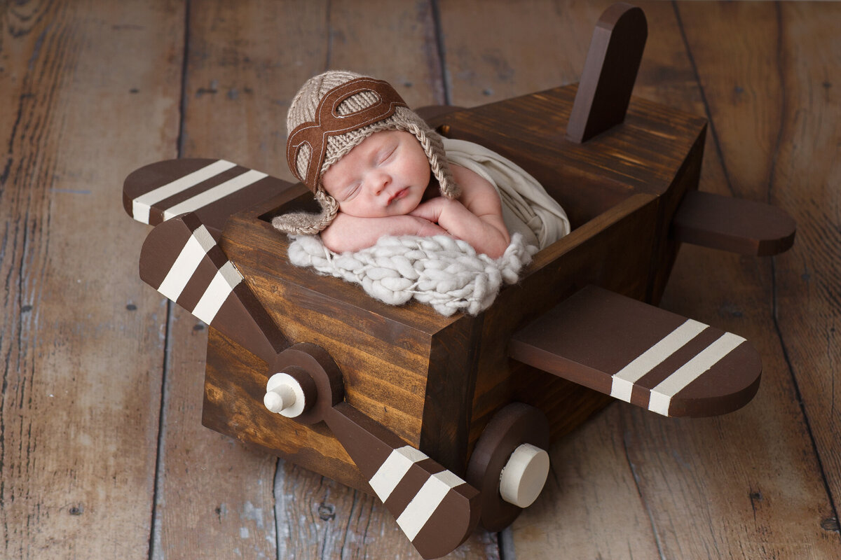Image of a newborn baby wearing an aviator cap and glasses and sitting inside a wooden airplane prop