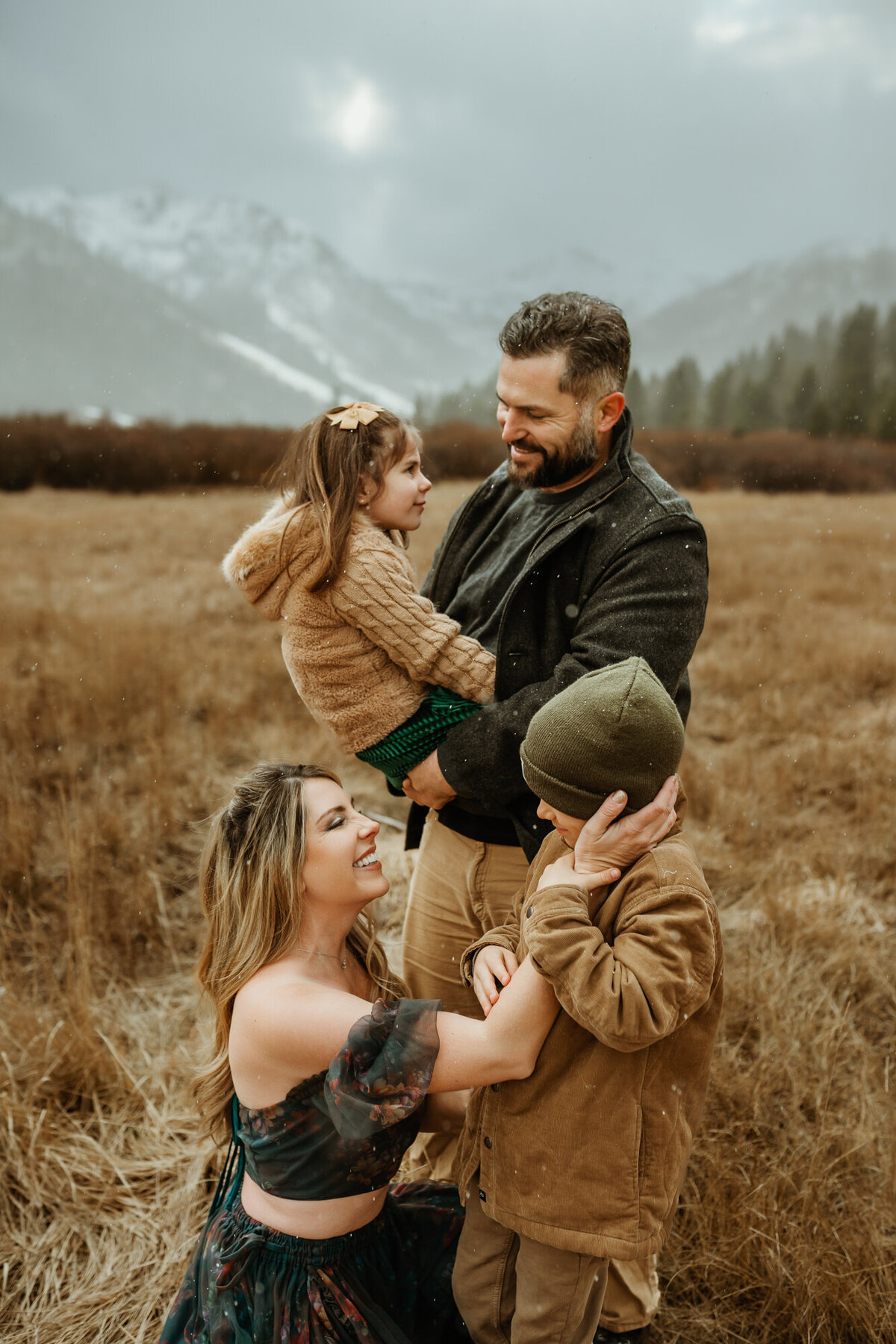 Mom is kneeling and holding her son's cheek, and the dad is behind mom standing while holding their daughter in a field with snowy mountains