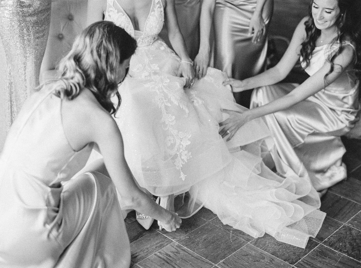 black and white portrait of bridesmaids helping bride put her shoes on in her wedding gown