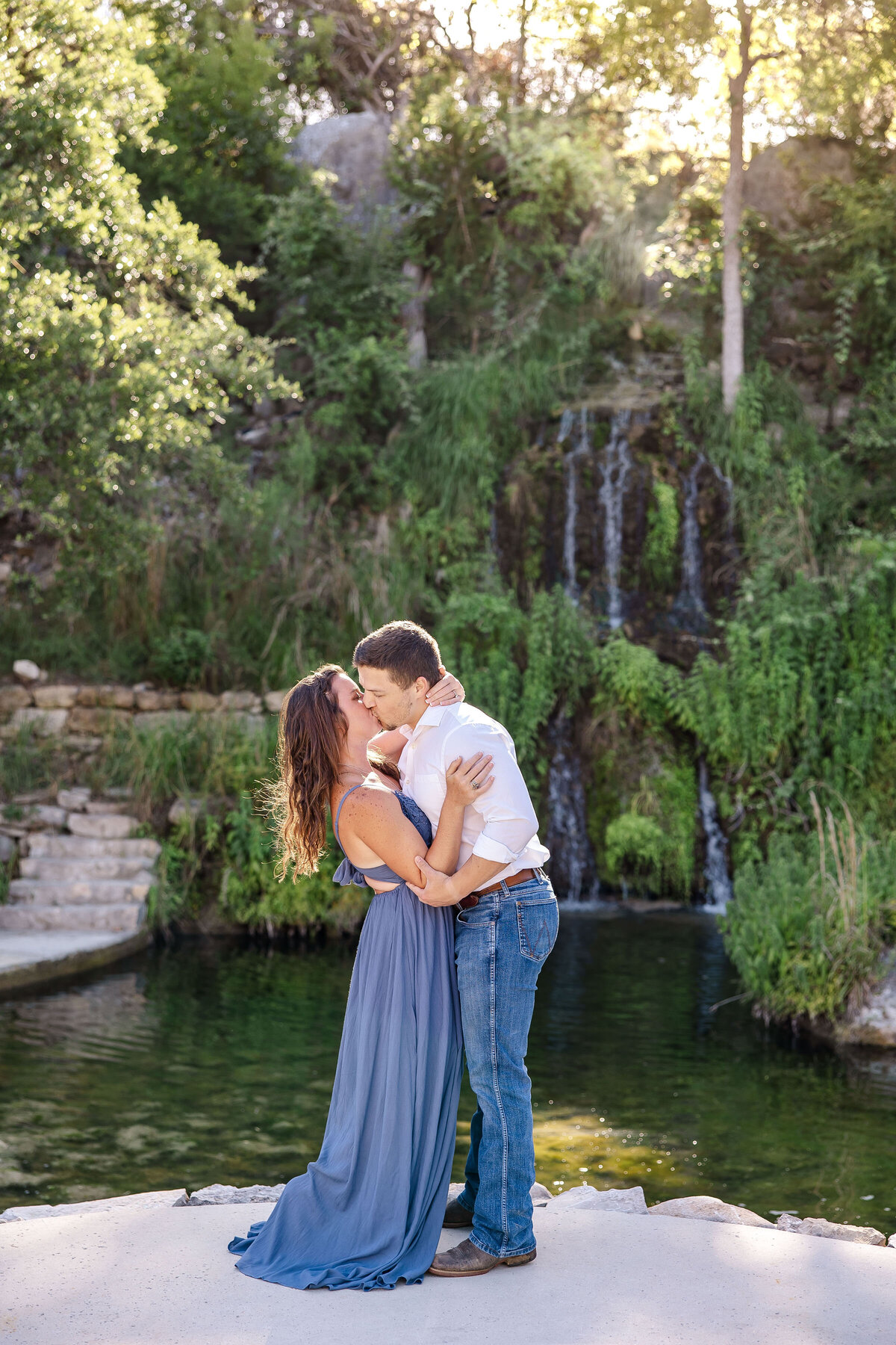 couple kiss  with waterfall and sun casting through trees at Remi's Ridge Hidden Falls in Spring Branch Texas