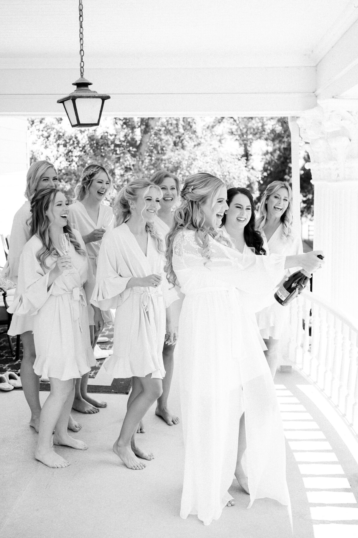 A group of women in robes celebrate on a porch at a modern and romantic wedding in Calgary, with one woman holding and opening a champagne bottle.