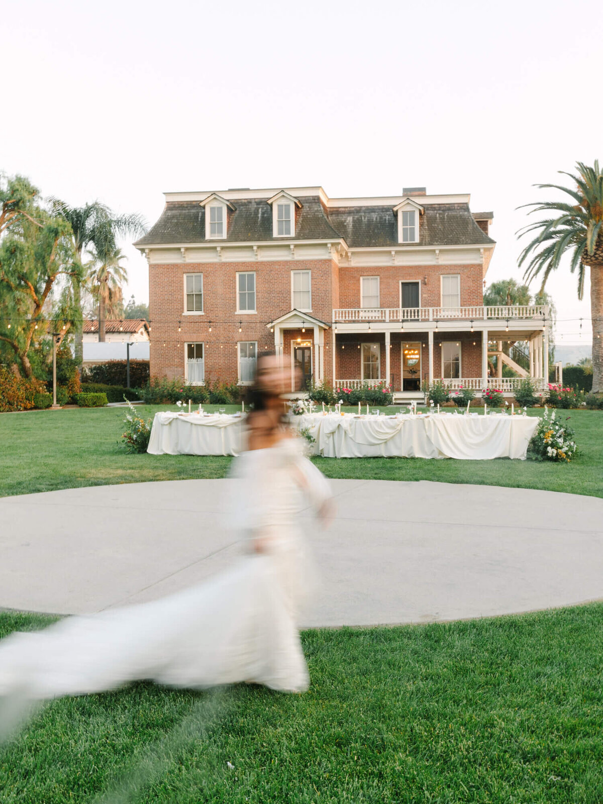 A blurred bride in a flowing white gown twirls joyfully in front of a large, elegant brick mansion.