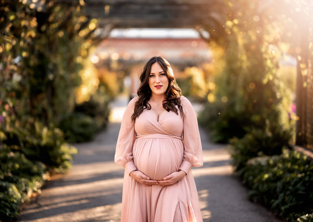 A beautiful young mama to be in a long pink dress stands in the sunlight  on a garden pathway