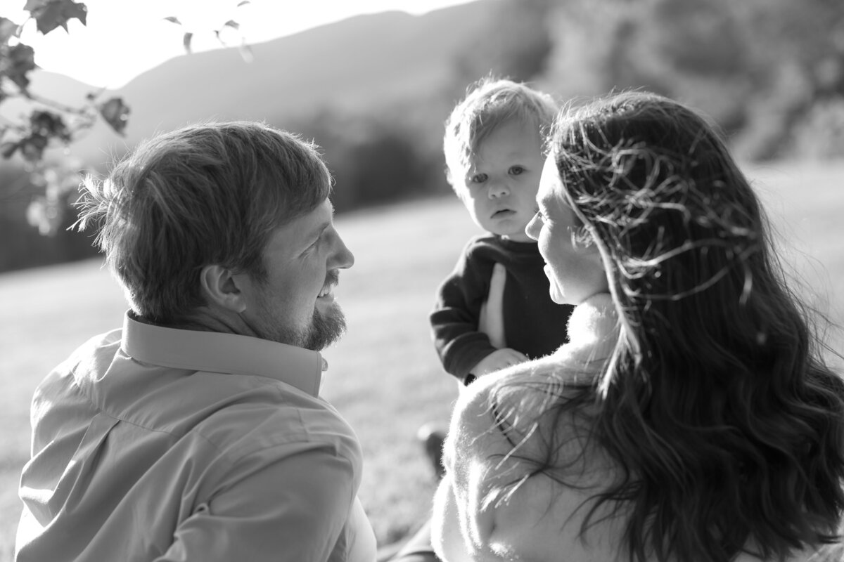 Black and white photo of a family outdoors. A couple sits with their toddler on a grassy New England field. The father looks at the child, who gazes at the mother. Sunlight softly illuminates the scene, with hills in the background—perfect for a New England photo book.