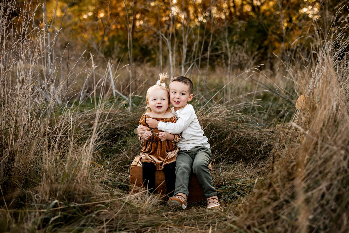 A young boy and girl sit on a wooden box in a grassy field. The boy, wearing a white shirt and green pants, has his arm around the girl, who wears a brown dress. They are surrounded by tall dry grasses with autumn-colored trees in the background.