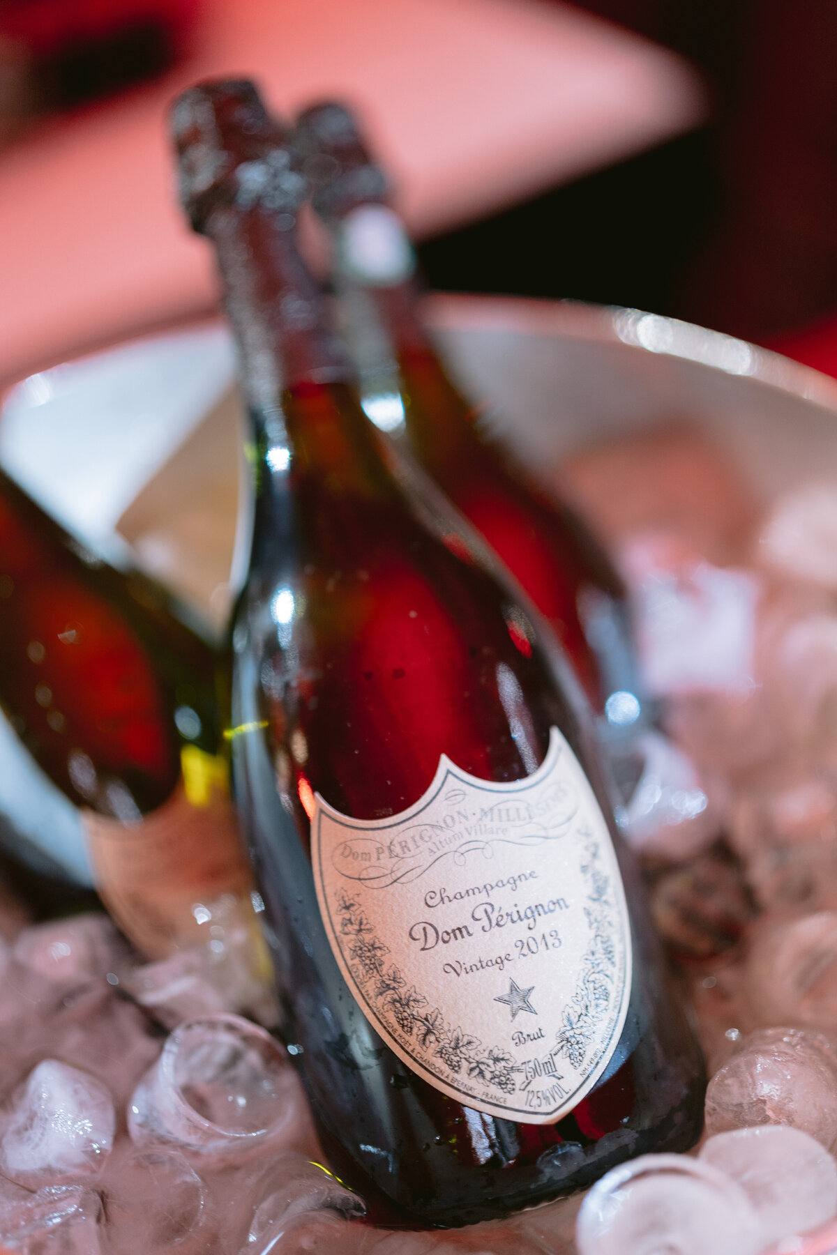 The photo was taken at Shangri La Hotel & Ritz Paris Hotel in Paris, France, by photographer Matteo Coltro and videographers Zen Film Works. In the image, two bottles of Dom Pérignon champagne are chilling in a bucket filled with ice cubes. The focus is on the front bottle, displaying its label indicating it's a vintage 2013. The scene suggests a festive or celebratory setting.