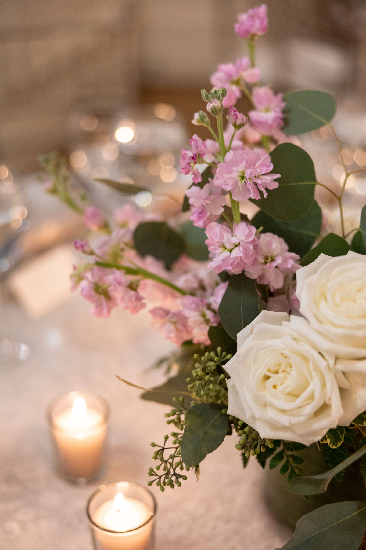 A floral arrangement with white roses and purple flowers, surrounded by green eucalyptus leaves, is placed on a table. Two small lit candles are near the bouquet, creating a warm and cozy ambiance.