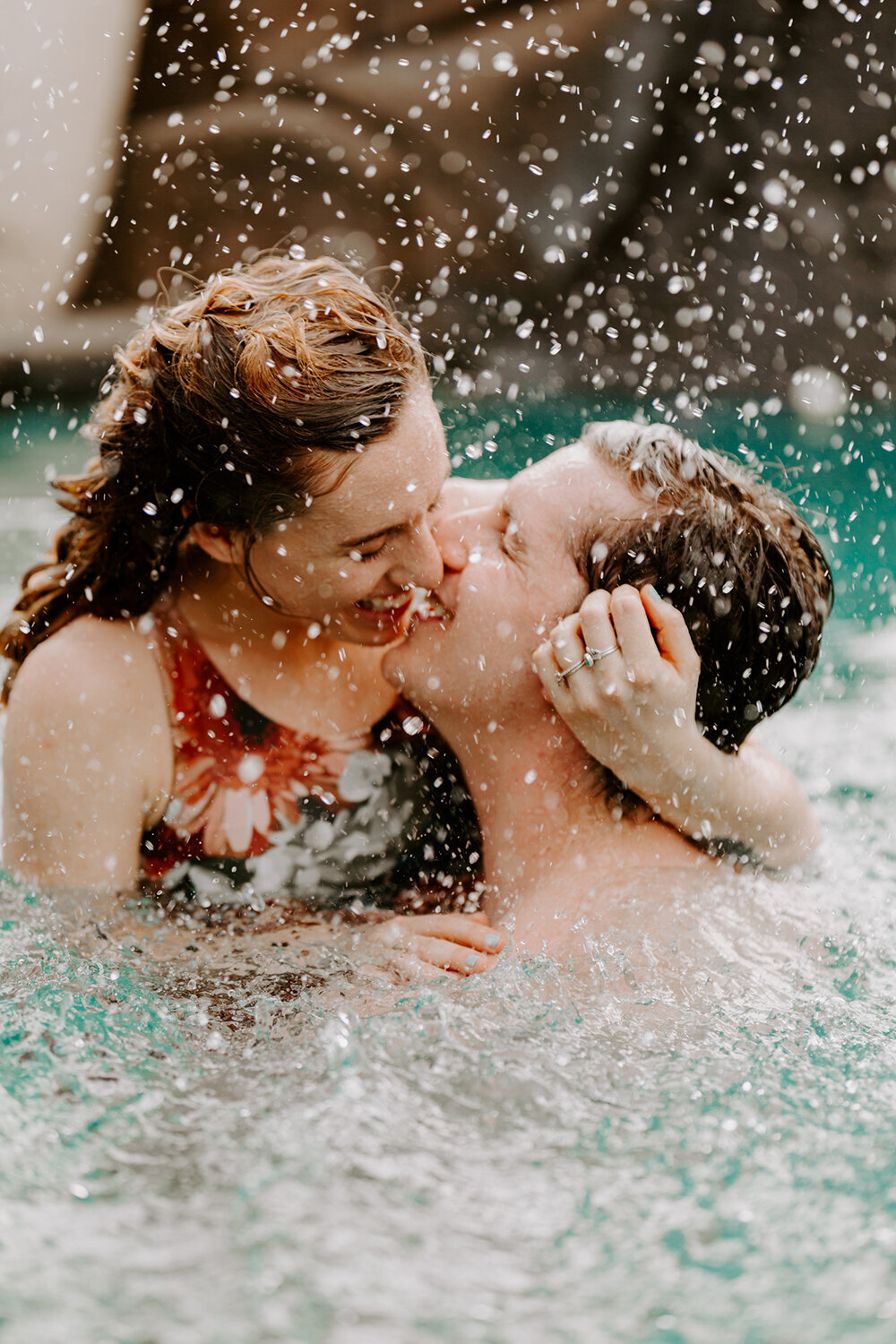 Cndid, adventurous couples sessions are so much fun! A young couple kiss and laugh and hold each other while splashing around in a pool in Hawaii.