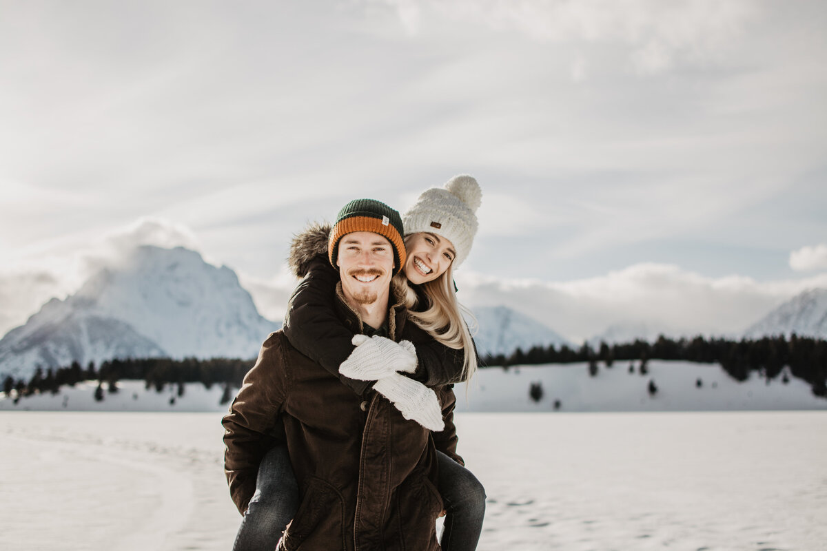 engaged couple in the Tetons during winter for an engagement session, blonde woman on his back holding onto his shoulders.