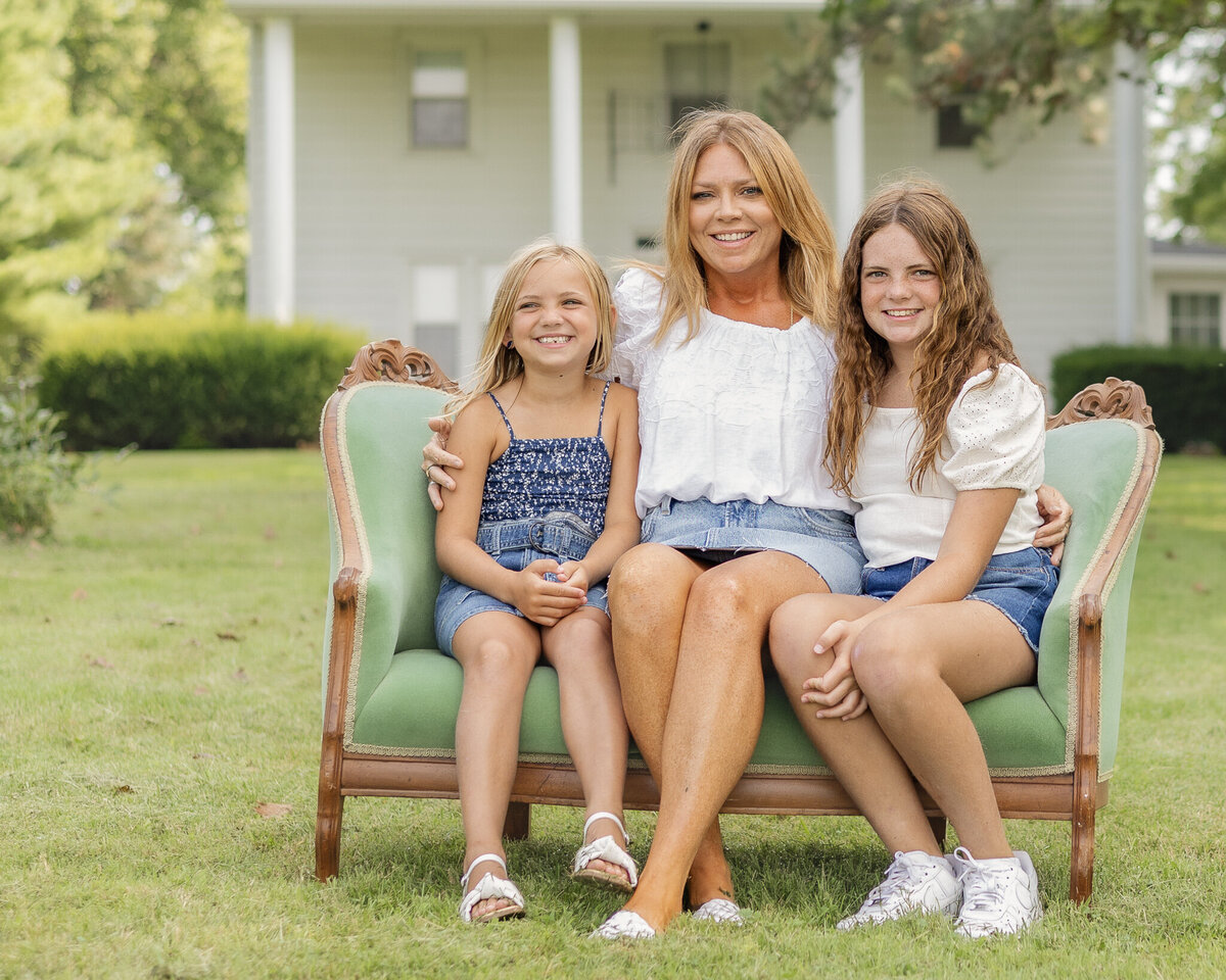 Mom and daughters are sitting in the vintage green couch in the grass with their family home in the background, everyone smiling at the camera.   Everyone is looking at the camera smiling.  Mom is wearing a jean skirt, white half sleeve shirt, and has long blonde hair.  Older daughter is wearing blue jean shorts, a white short sleeve shirt, and has long brown hair.  Youngest daughter has a dark blue and white tank top style shirt with a blue jean skirt, and has medium length blonde hair.