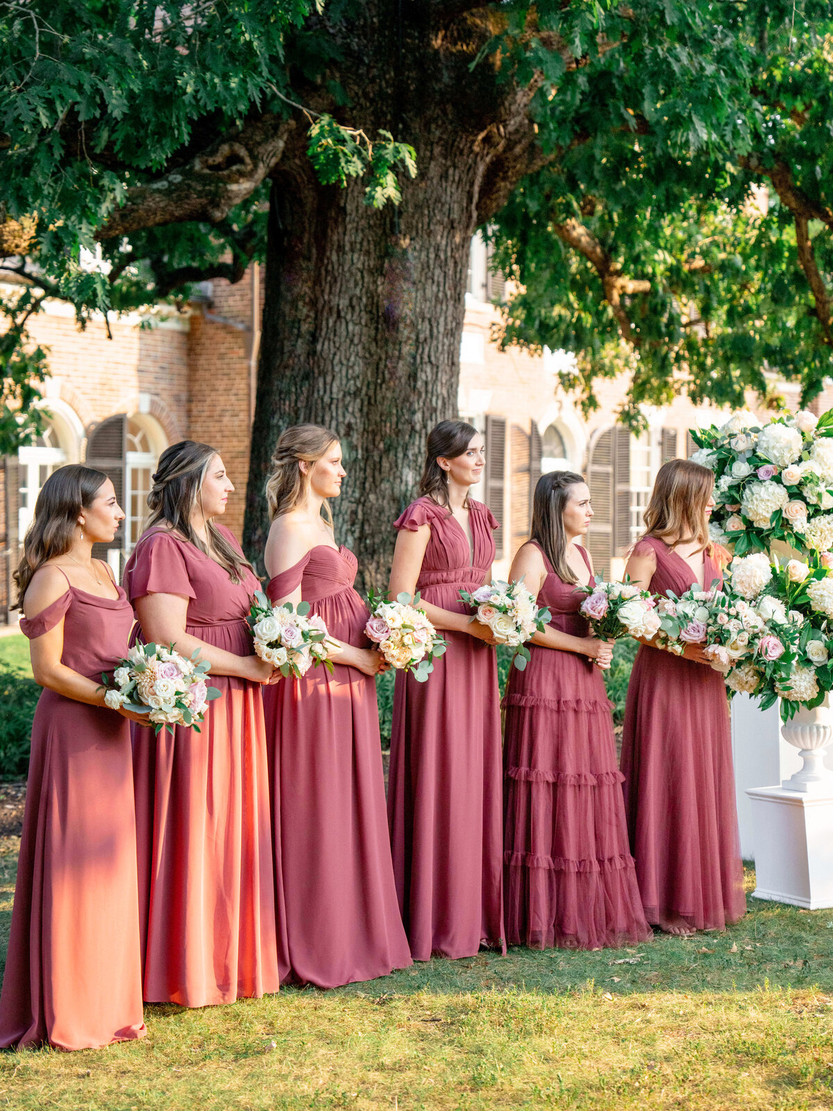 A group of bridesmaids stands outdoors in matching burgundy dresses, holding bouquets of pastel flowers. They are positioned under a large tree, near a brick building. A large floral arrangement is visible to their right.