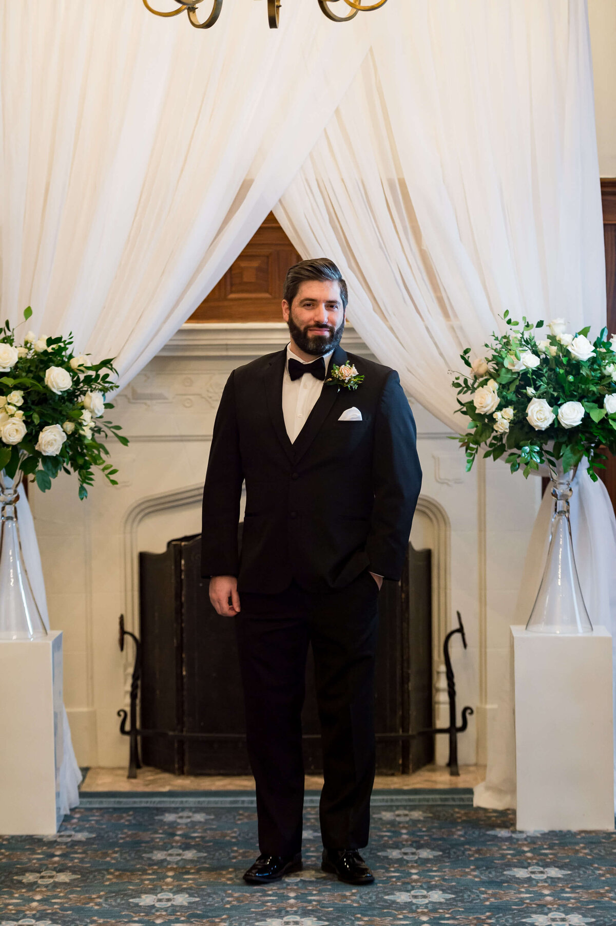 a groom in a black tuxedo is waiting at the altar for his Ottawa wedding at the Chateau Laurier hotel to start.  His hand is in his pocket and there is elegant draping and a fireplace in the background.