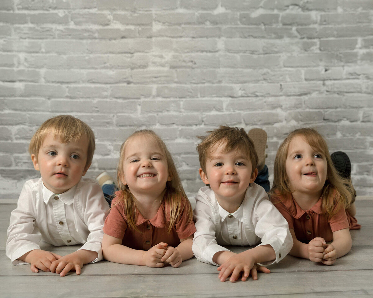 Quadruplets laying on their stomachs in front of a grey brick background.