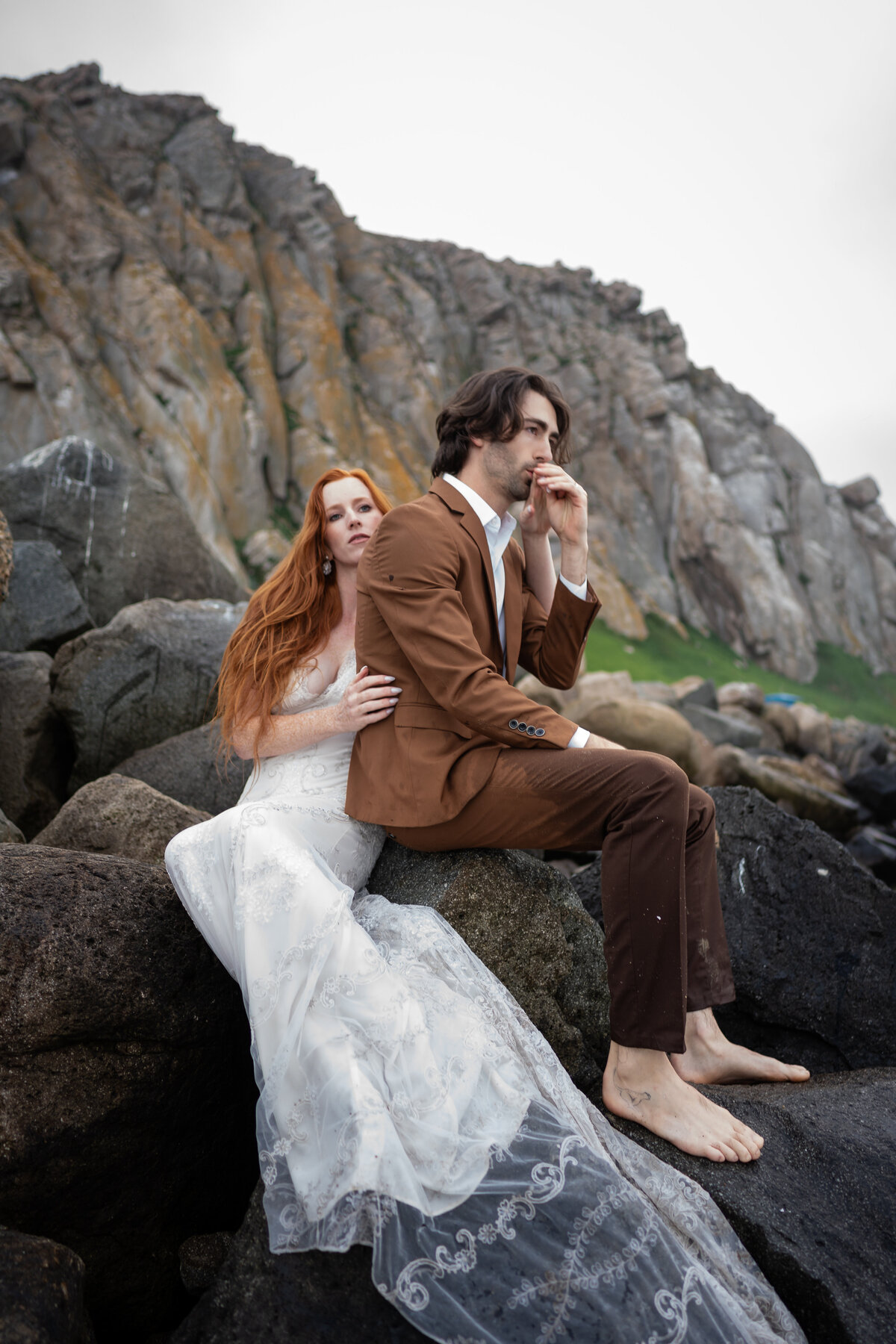 Bride and groom at Morro Bay coastal California sitting barefoot on rocks embracing each other with a Claire Pettibone lace gown