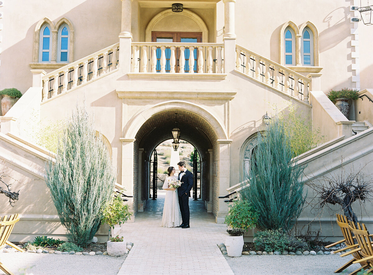 Kohnur Elegant couple portrait with bride in natural makeup and a romantic updo wearing a flowy A-line gown at the base of a grand European chateau staircase.