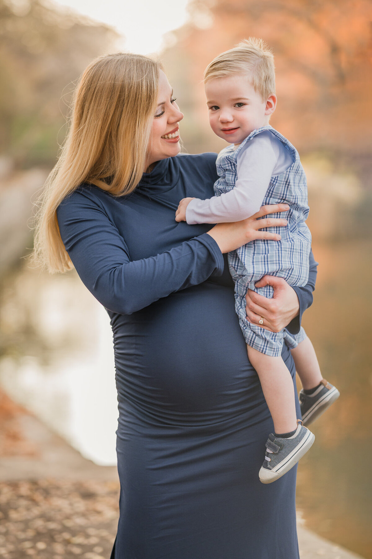 Pregnant woman holds her toddler and smiles at him during maternity pictures at Brackenridge Park in San Antonio.