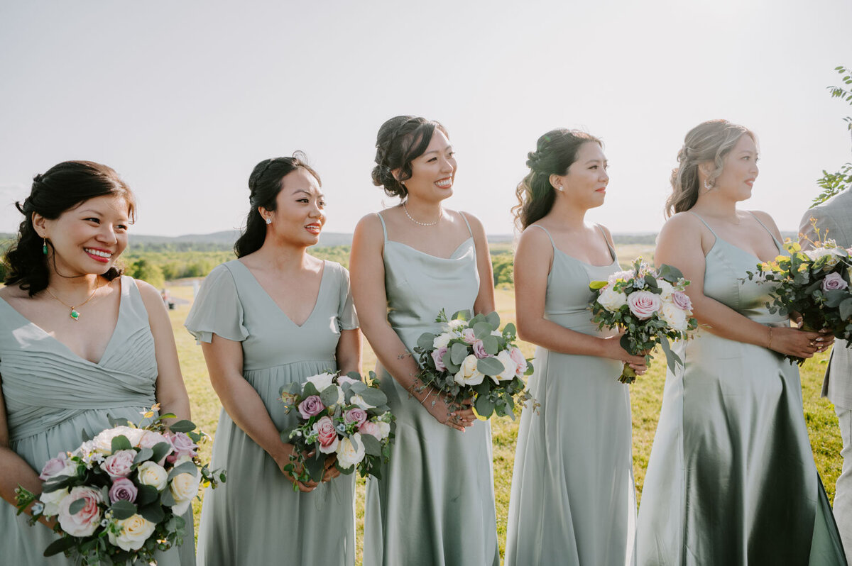 Bridesmaids standing outdoors in sage green dresses, holding bouquets and smiling.