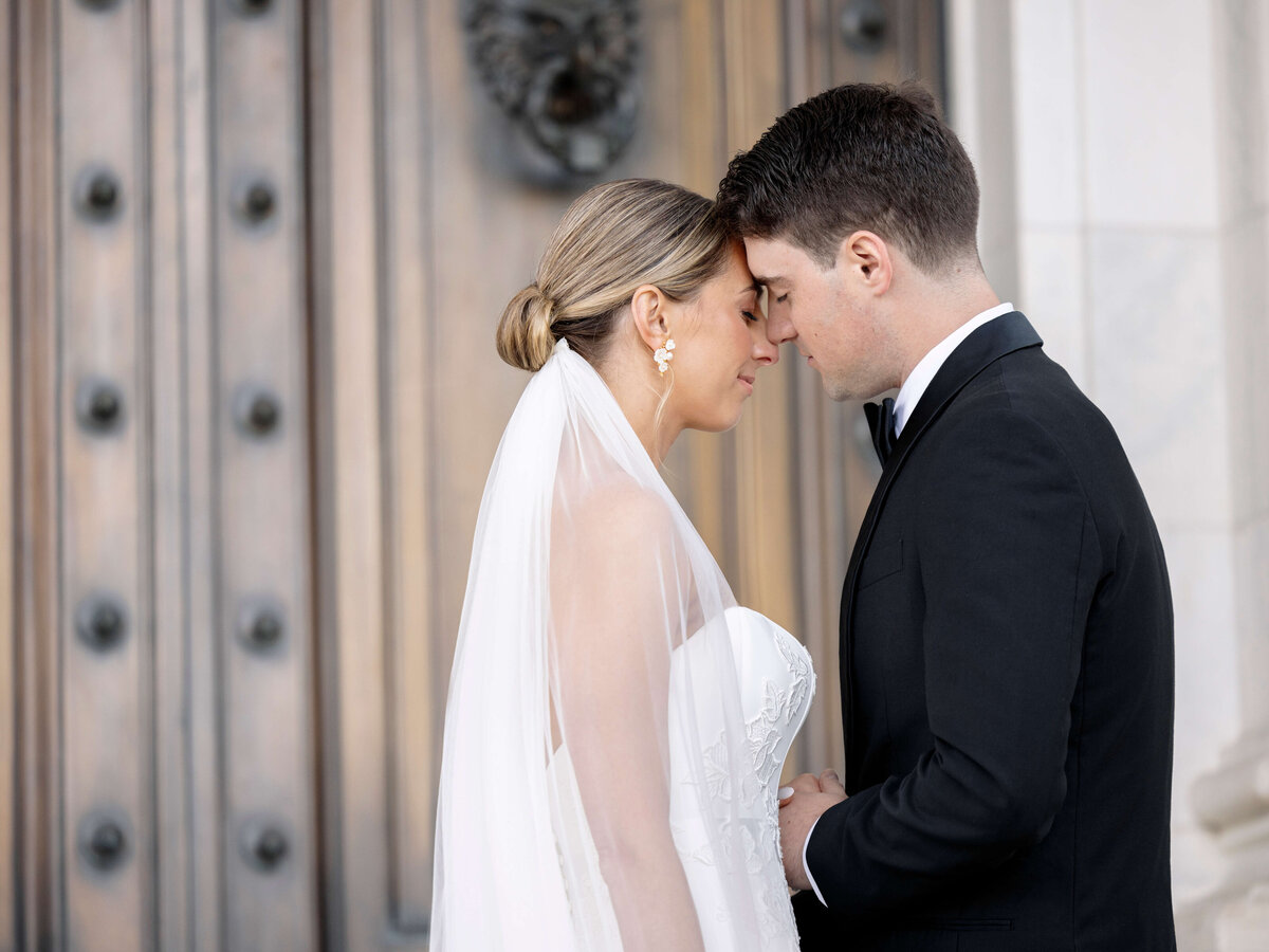 A bride and groom stand with their foreheads touching in front of a large ornate door. The bride wears a strapless white dress and veil, while the groom is in a black suit. They hold hands gently, creating an intimate and serene moment.