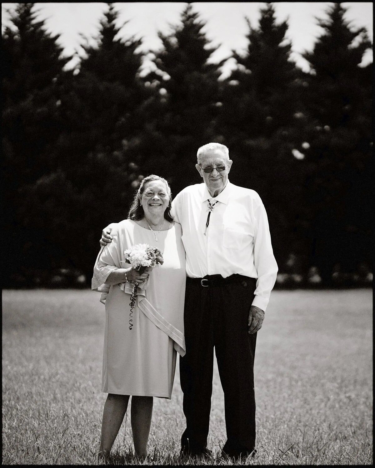 Elderly couple standing arm-in-arm in a field, with trees in the background