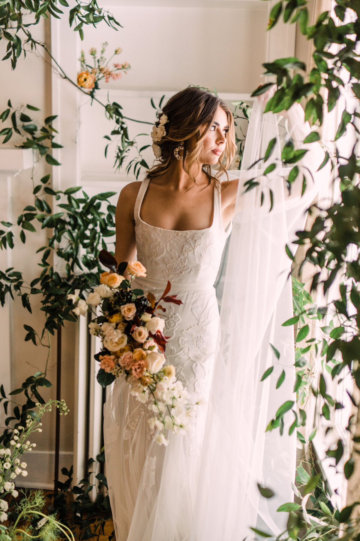 Bride wearing Alexandra Grecco Marcelle Gown holding bouquet in front of vine-covered window at Cornman Farms