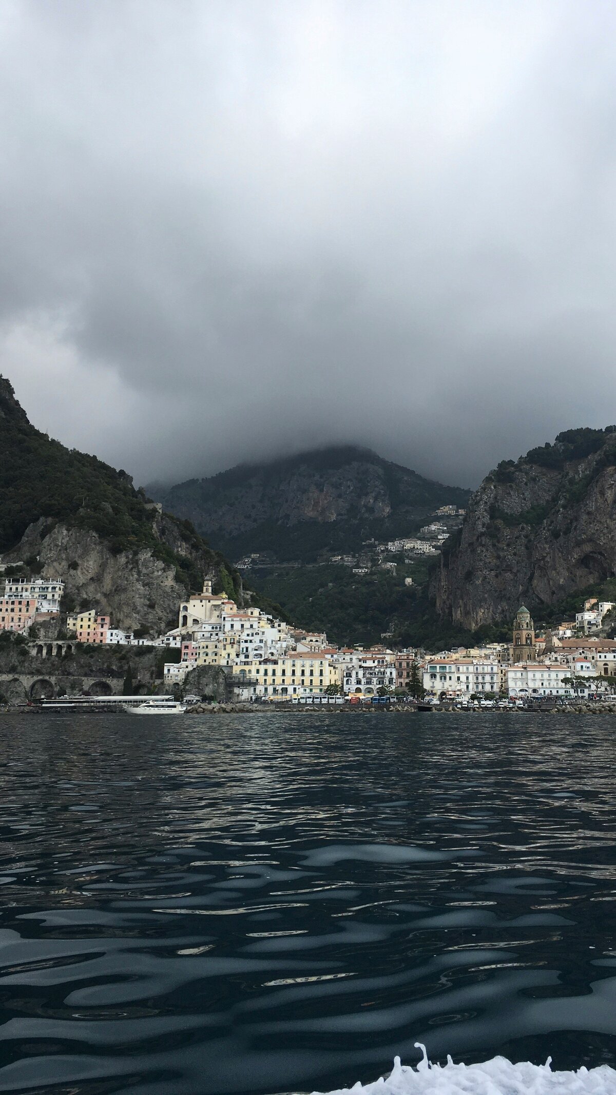 Picturesque coastal town in the Amalfi Coast, Italy, nestled below dramatic cloudy mountains, hinting at the scenic diversity of the Velaris Escape literary tour.
