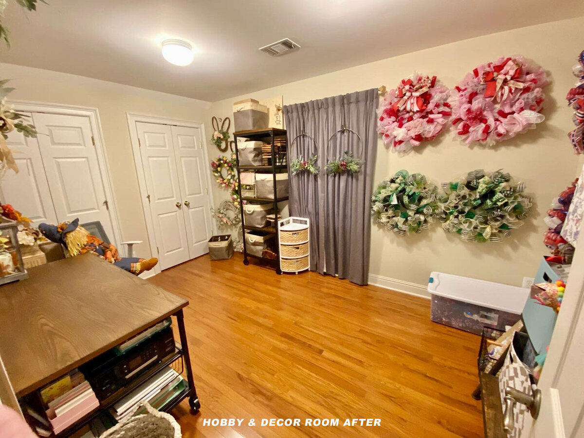 A well-organized hobby and decor room with wooden flooring. It features a workstation with a table, storage shelves, and various floral wreaths decorating the walls. A basket rack and plastic storage bins are neatly arranged.