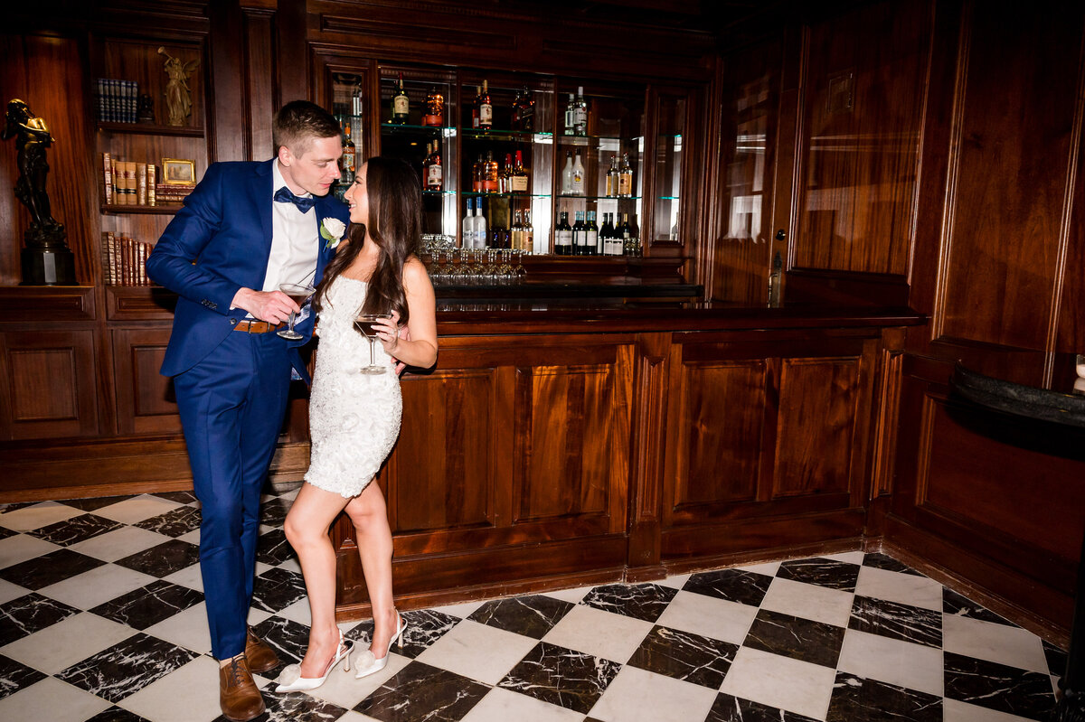 A man and woman stand near a wooden bar, both holding drinks. The man wears a blue suit, and the woman is in a white dress with high heels on a checkered black and white floor.