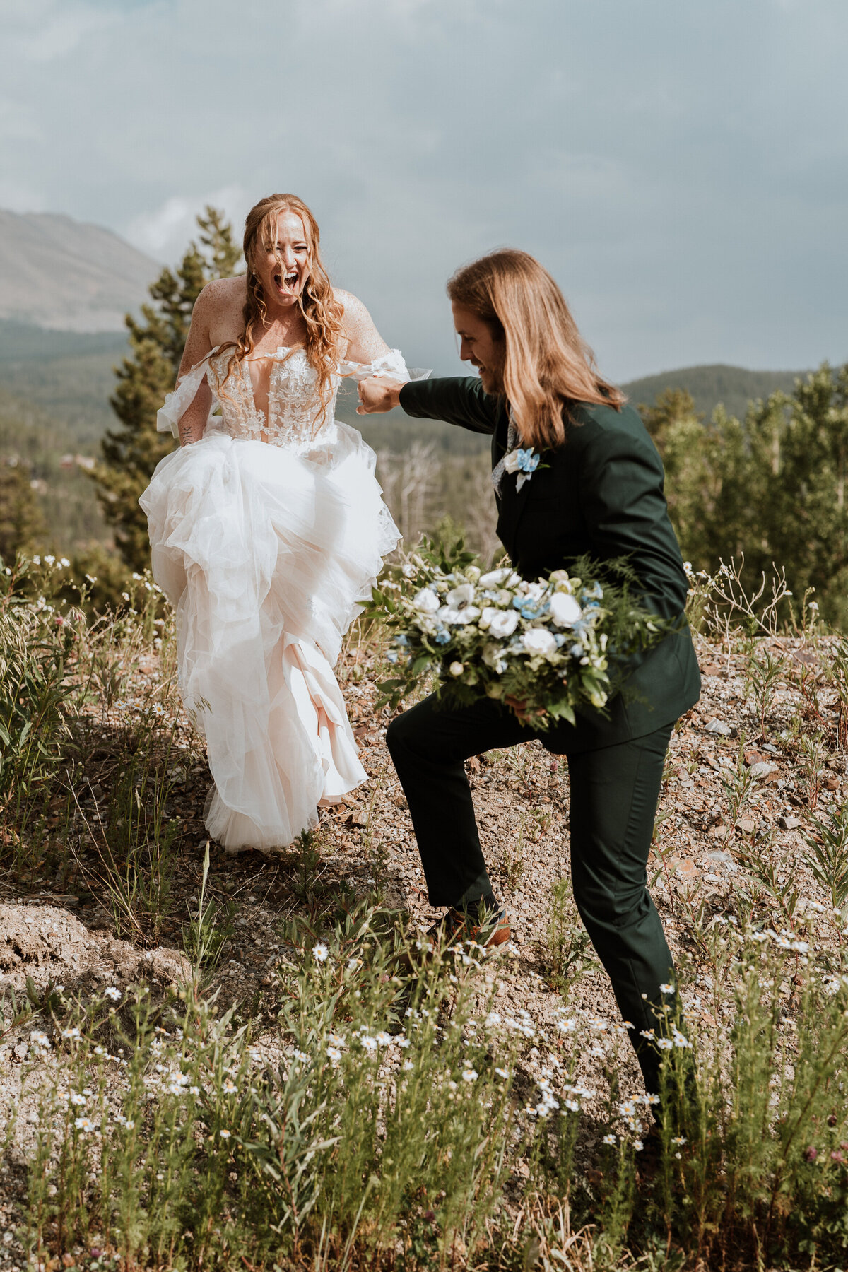 Bride laughs while partner helps her up a hillside during adventurous Breckenridge mountain wedding.