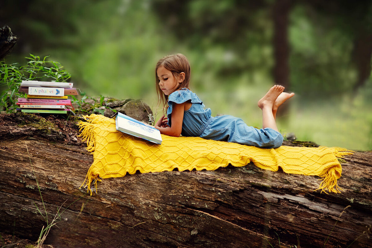 Little girl lays on her belly on a blanket on a big log in the forest while reading a book. Captured in 100 Mile House by Brittany Danielle Photographu=y