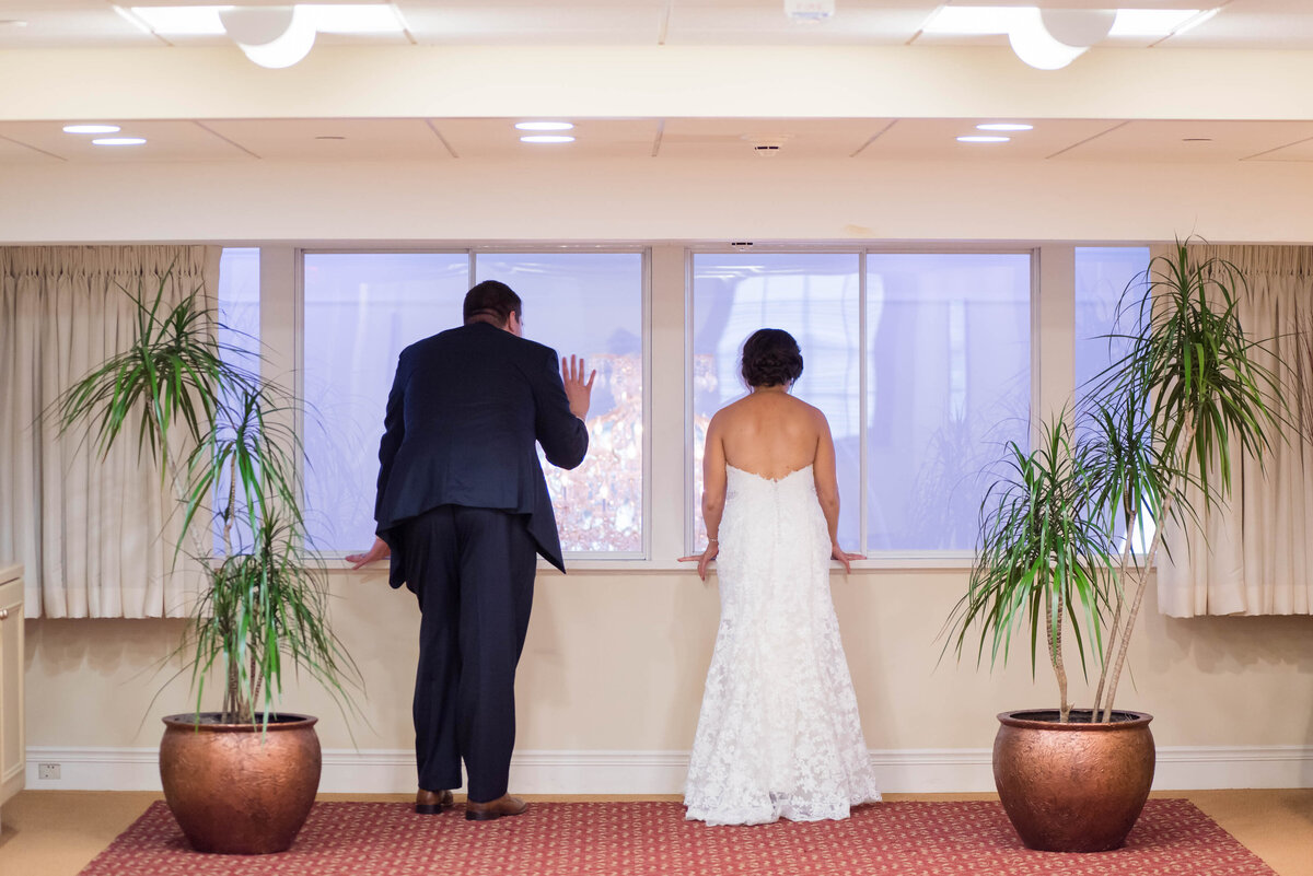 bride and groom peeking through the windows at university club overlooking their wedding reception as guests enter