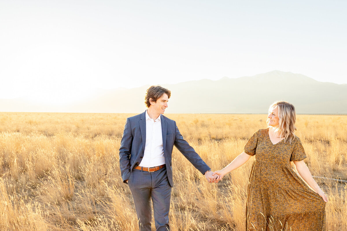 A couple walks hand in hand through a field of golden grass, smiling at each other with mountains in the distance.