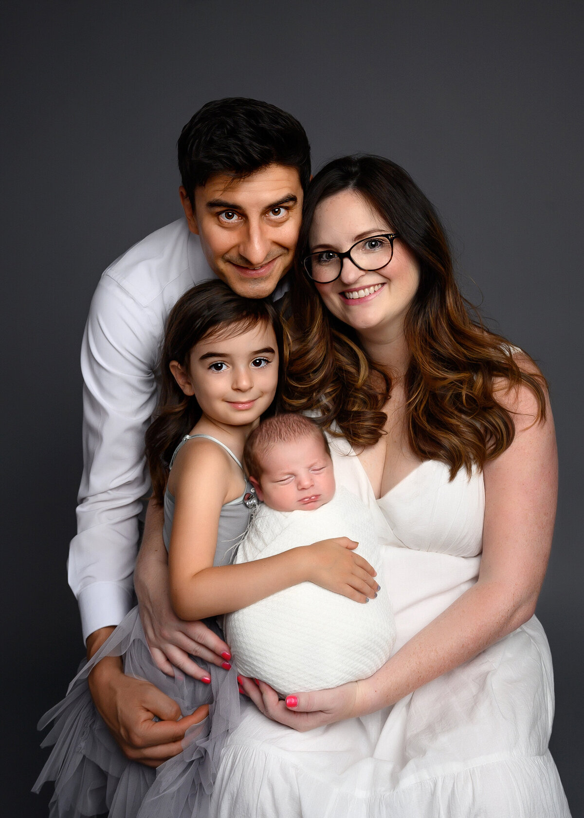 studio session of a family with a new addition, father embracing mom and children on a gray background